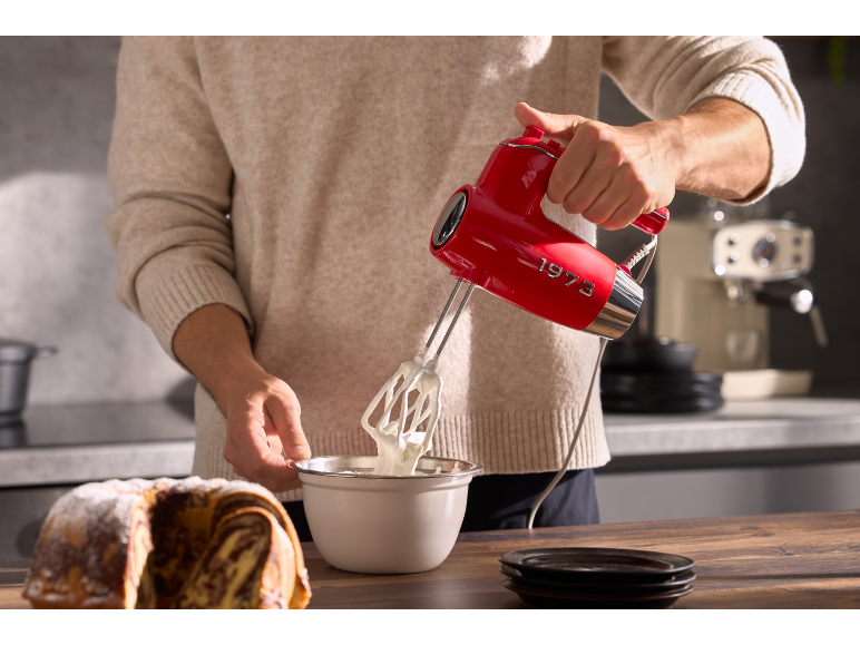 Man whisking cream with a red hand mixer, with a cake beside it.