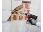 A cordless rotary hammer with a chisel removes tiles from a wall, revealing brickwork.