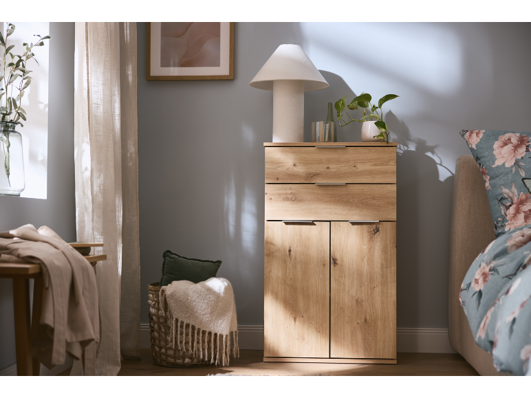Wooden cabinet with drawers and doors, table lamp, plant, and basket with blanket in a bedroom.