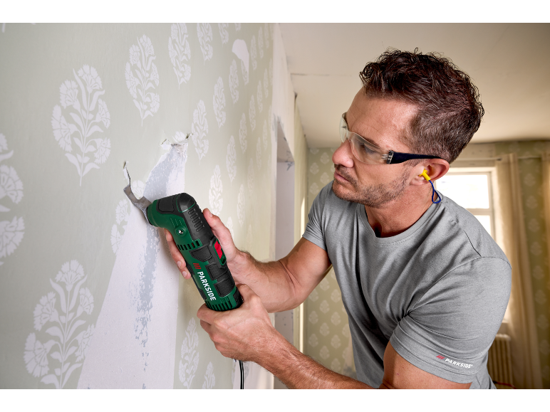 Man using a multi-tool to remove wallpaper, wearing safety glasses and earplugs.