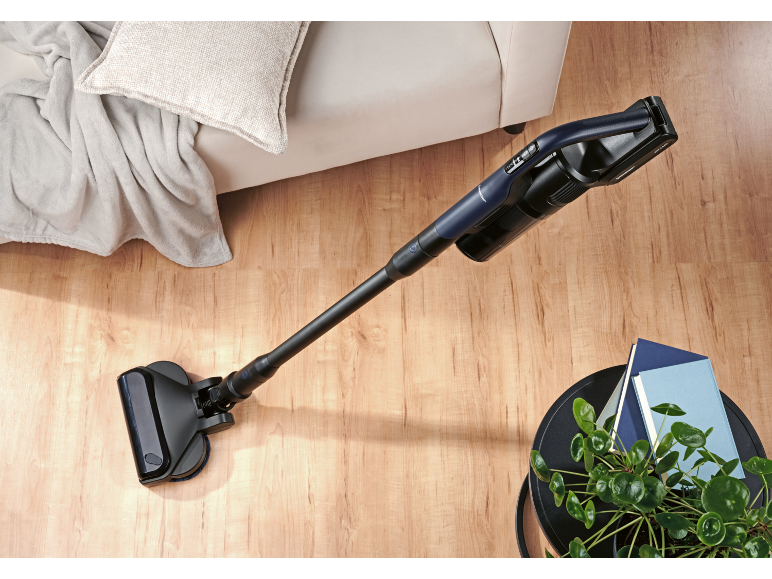 Blue cordless vacuum cleaner on wooden floor, with sofa and plant.