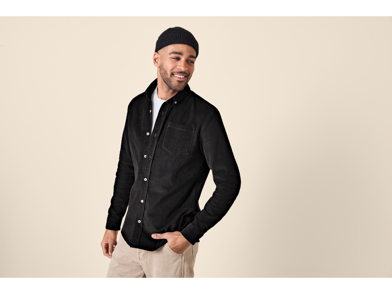 Man in black corduroy shirt and beige trousers, smiling and looking away.