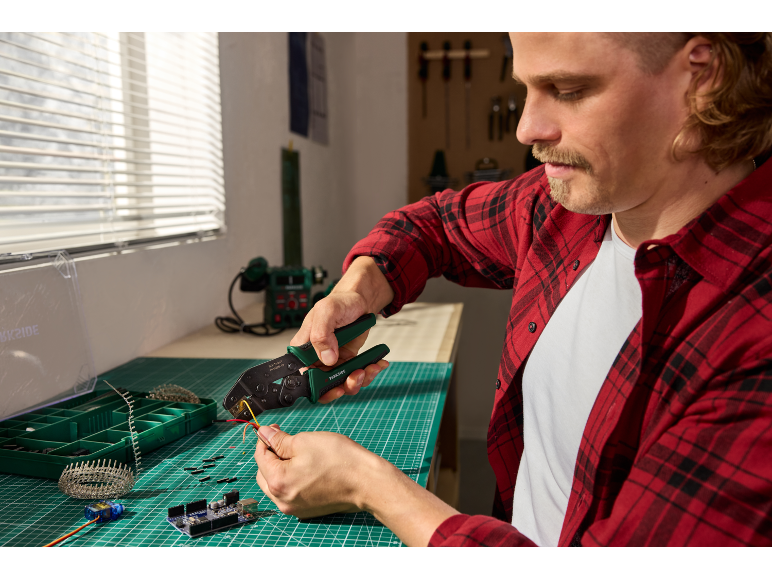 Man with crimping pliers and wires working on an electronics project on a green mat.