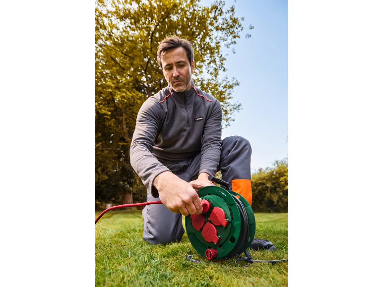 Man kneeling on grass, plugging a red cable into a green extension reel with red sockets.
