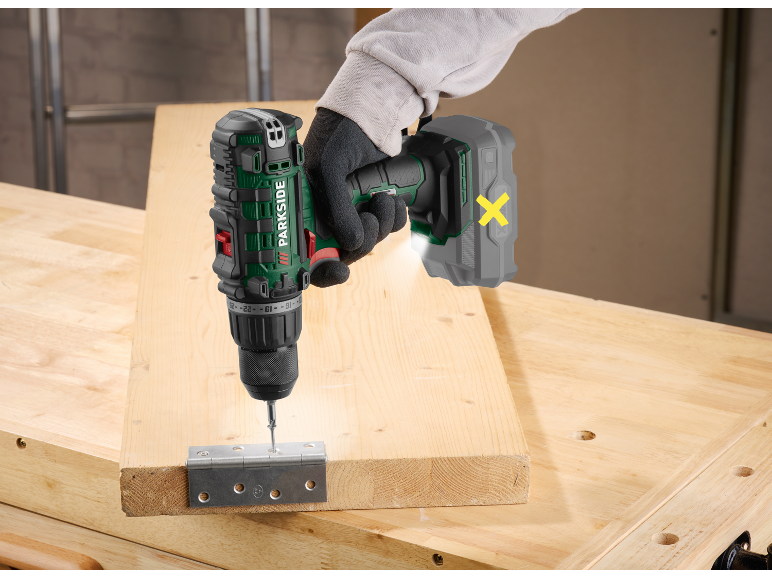 Gloved worker using a cordless drill to attach a hinge to a wooden board.