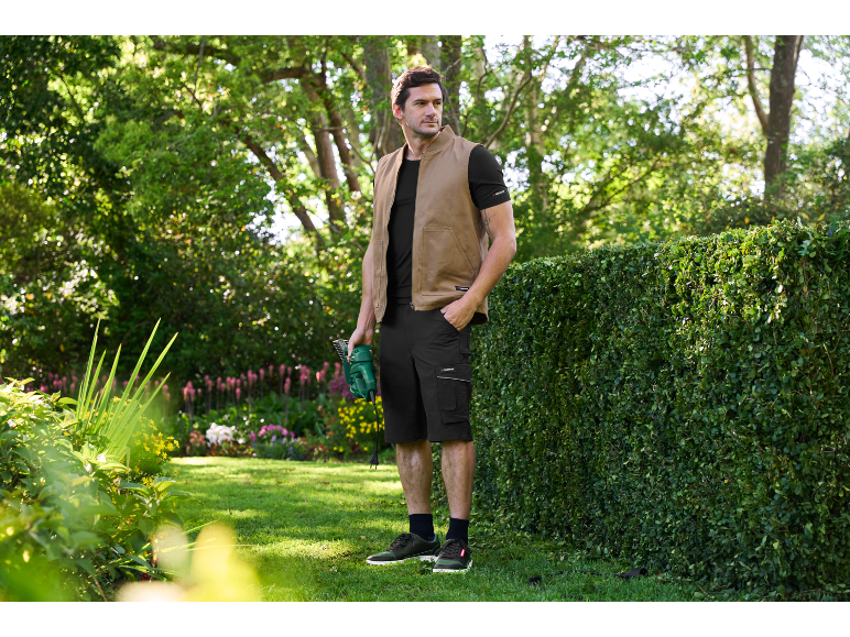 Man in a vest and shorts holding a hedge trimmer in a garden.