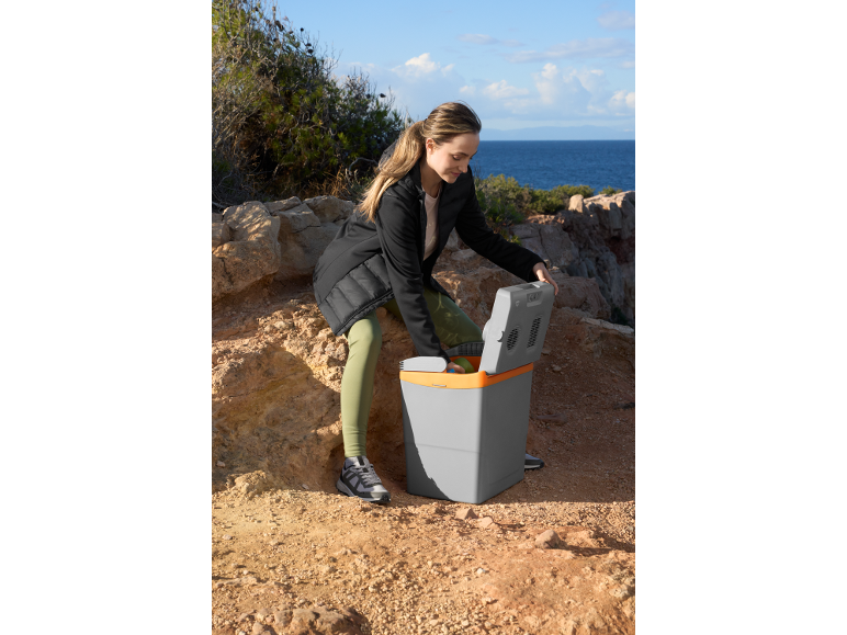 Woman opening a portable electric cool box on a rocky coastal path.