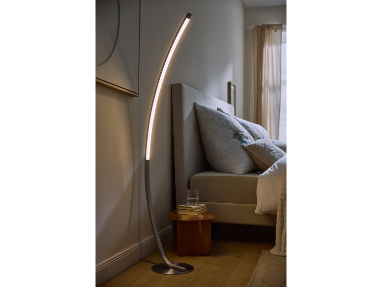 Curved LED floor lamp in a modern bedroom with a bed and nightstand.