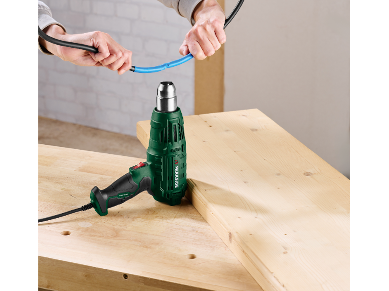 Hands holding a blue heat shrink tube over a heat gun on a wooden workbench.