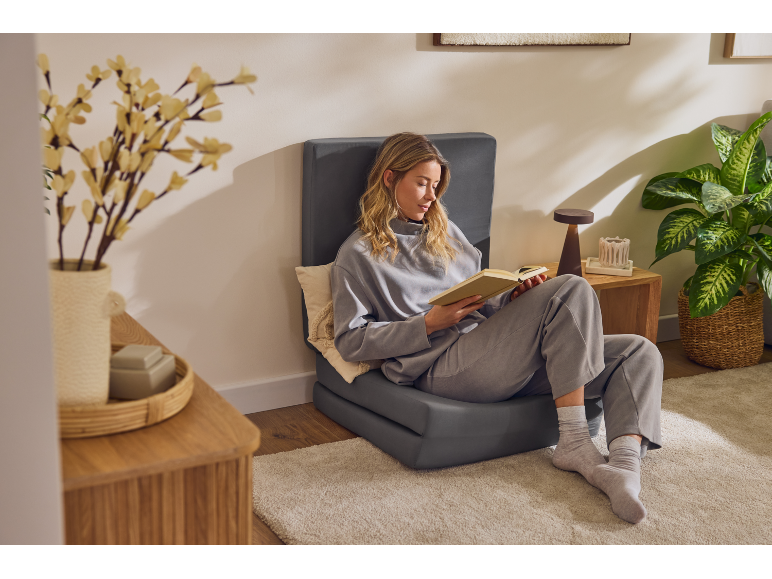 Woman reading a book on a grey floor seat, with a plant and a vase of flowers.