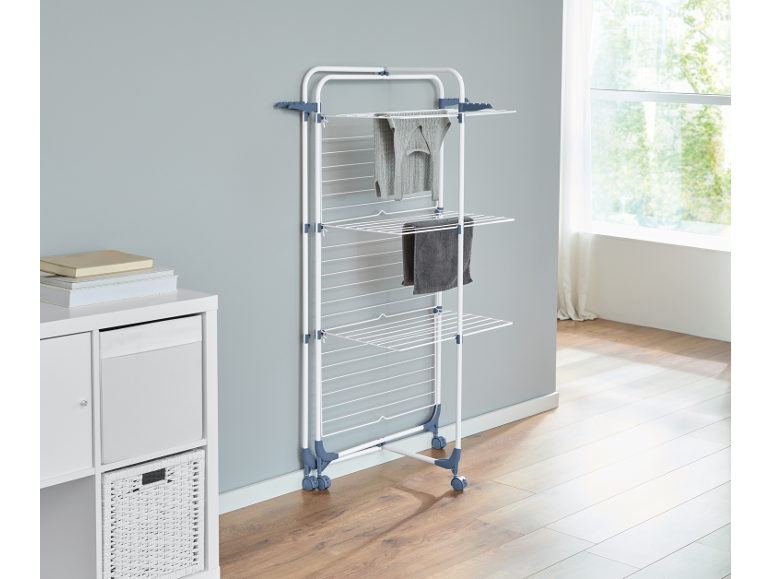 Vertical clothes drying rack with wheels, with clothes hanging, next to a white cabinet.