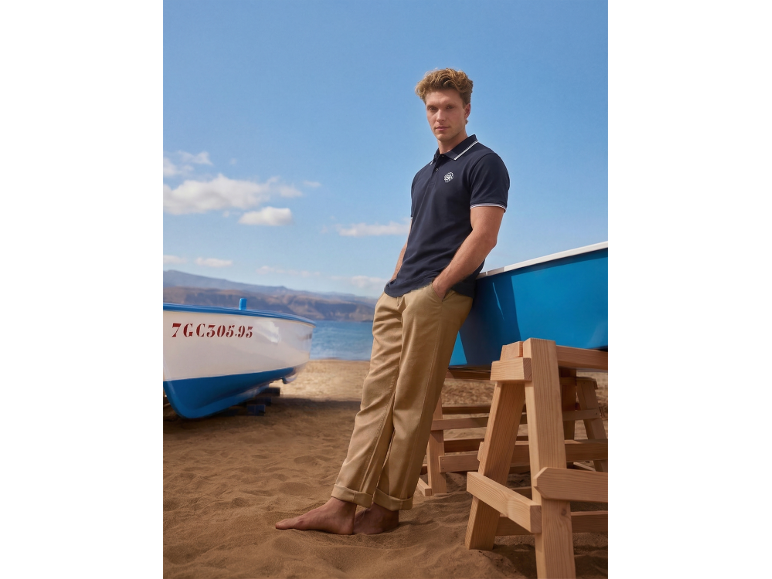 Man in navy polo shirt and khaki trousers on the beach, leaning against a boat.