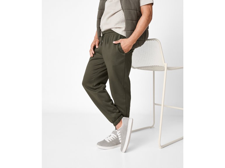 Man in olive green joggers, beige t-shirt, and quilted vest, leaning on a chair.