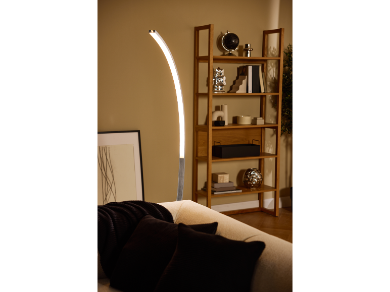 Curved LED floor lamp illuminating a sofa with cushions and a wooden bookshelf.