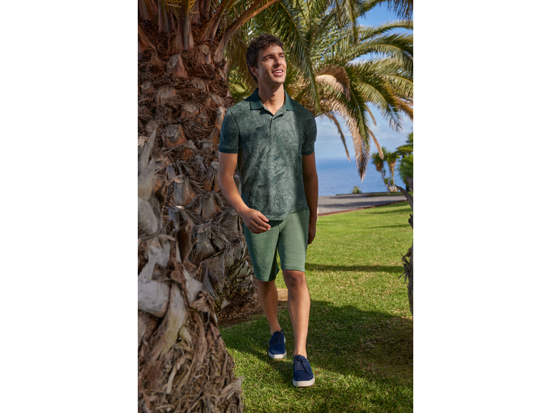 Man in green patterned polo shirt and shorts, walking among palm trees by the sea.