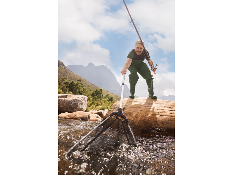 Man in waders with a fishing rod and net in a river.