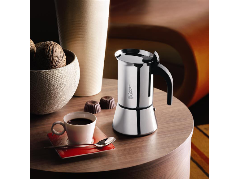 Stainless steel moka pot on a wooden side table with a cup of coffee.