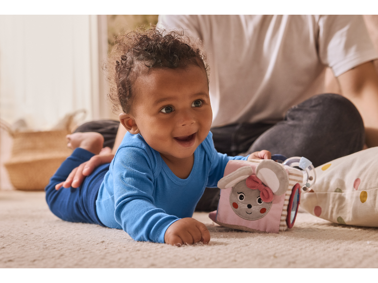 Happy baby in blue romper playing with a soft activity cube on the carpet.