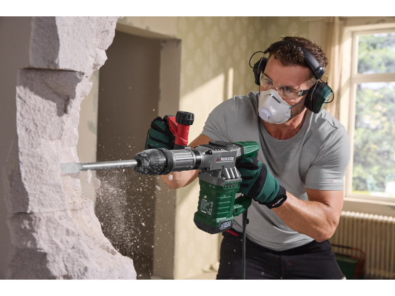 Man with a hammer drill, safety glasses, mask, and ear protection breaking down a wall.