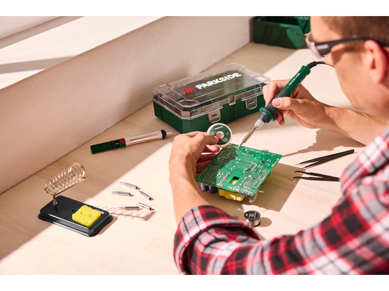 Man soldering a circuit board with a soldering iron, solder wire, and other tools on a workbench.