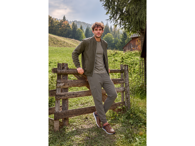 Man in khaki bomber jacket, T-shirt, and cargo pants, leaning on a wooden fence.