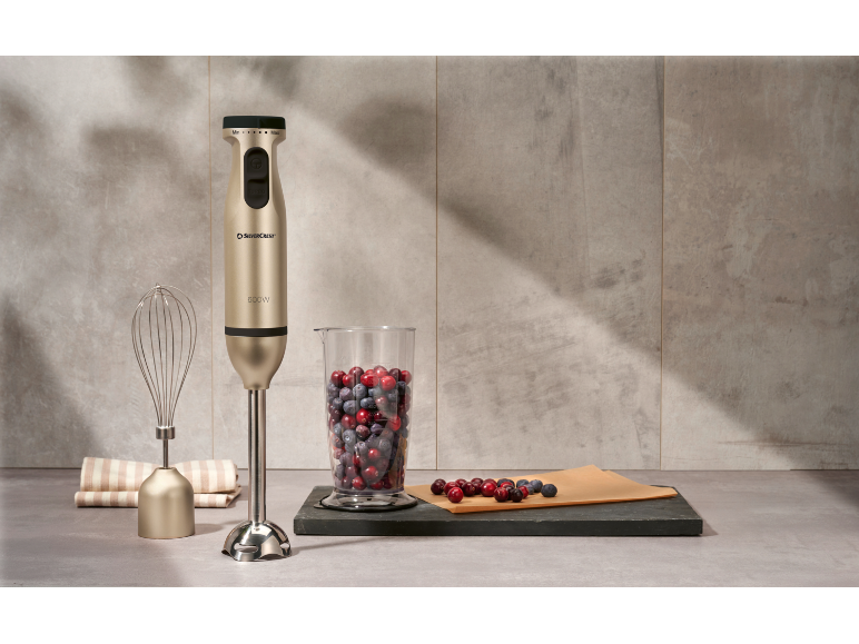 Gold immersion blender with whisk, measuring cup with berries, and kitchen towel.