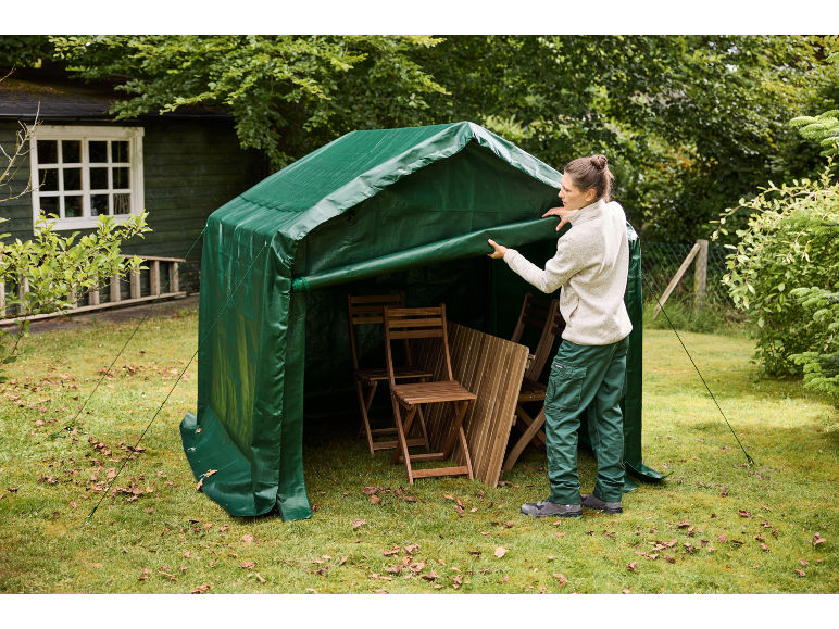 Woman closing a green garden tent with stored wooden garden furniture.