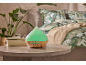 Green aroma diffuser with a wooden base in a bedroom with plants and patterned bedding.