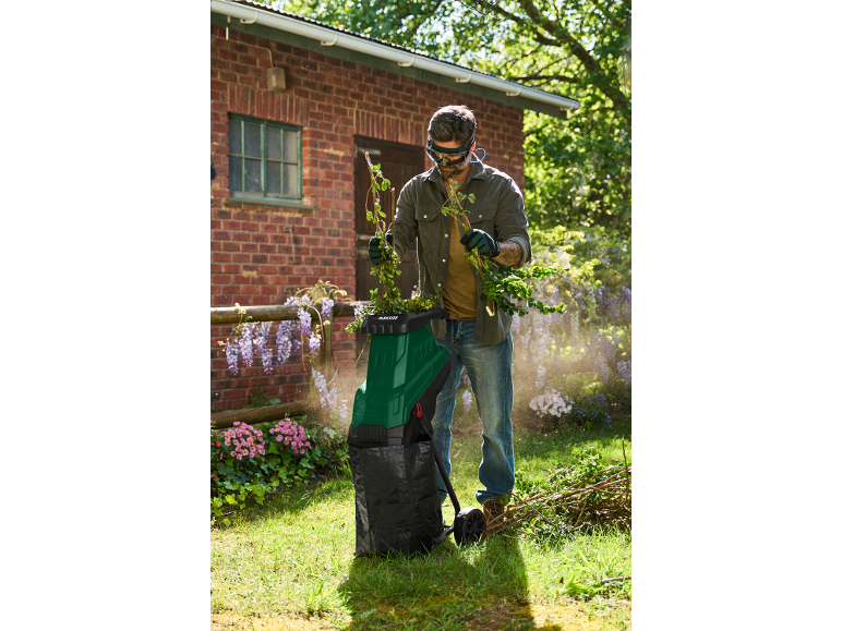 Man in safety glasses and gloves using a green garden shredder for branches.
