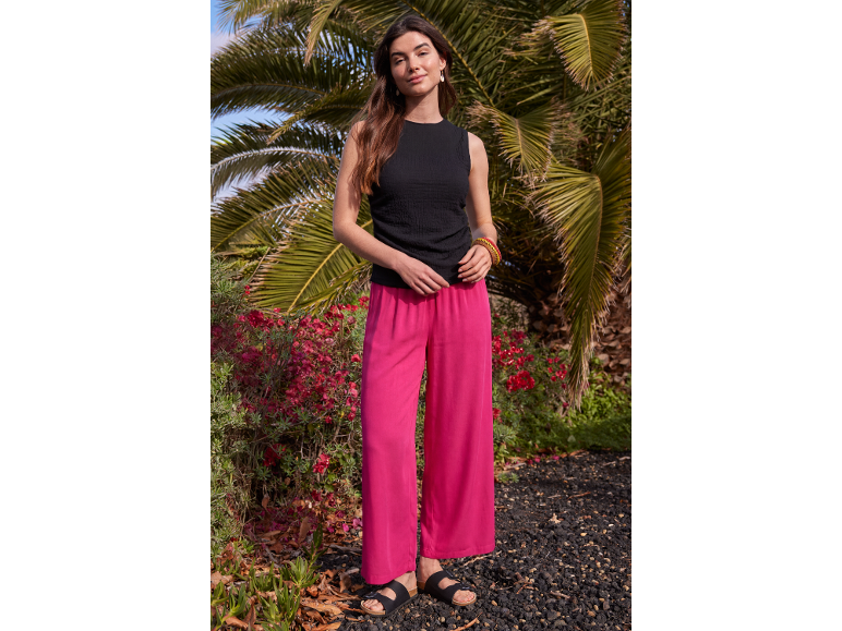 Woman in black top and bright pink wide-leg trousers in front of palm trees.