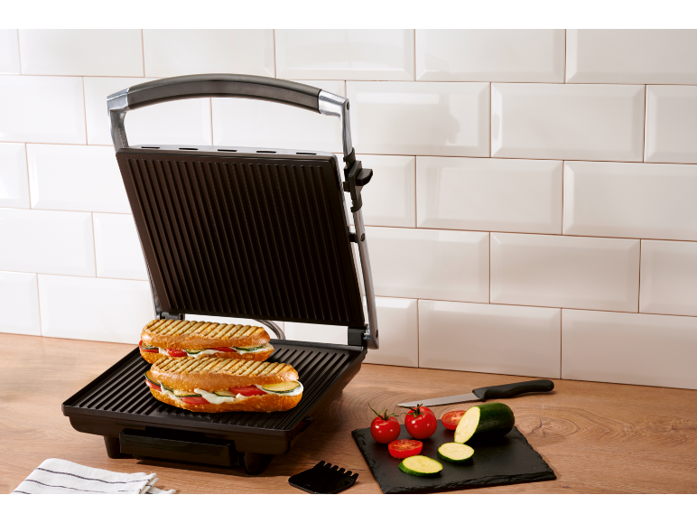 Sandwich maker with two grilled sandwiches and fresh vegetables in a kitchen.