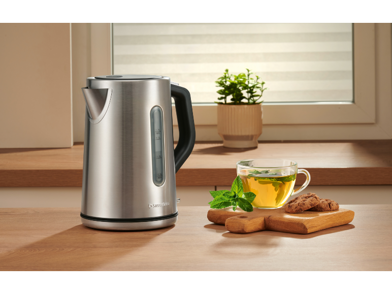 Silver kettle, teacup with mint, and two cookies on a wooden cutting board.