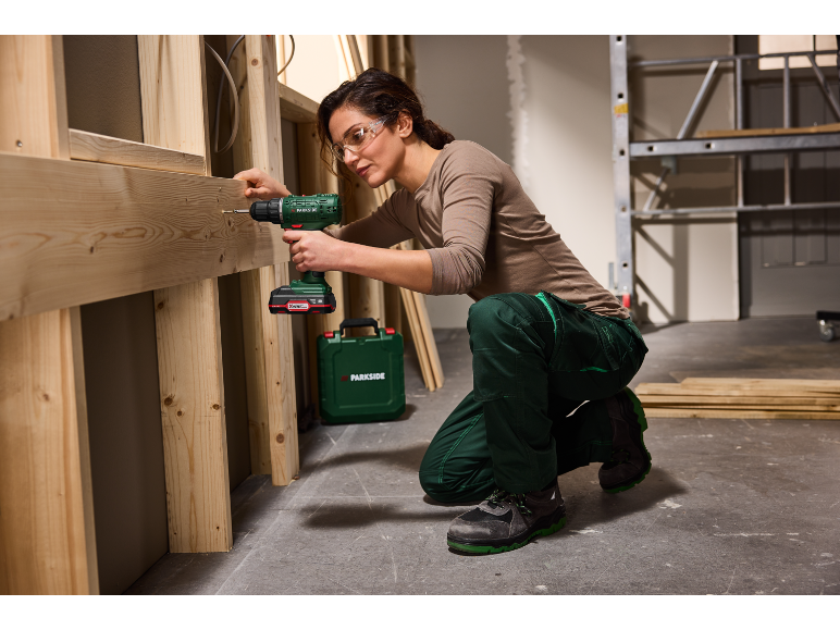 Woman in safety glasses using a cordless drill on a wooden construction.