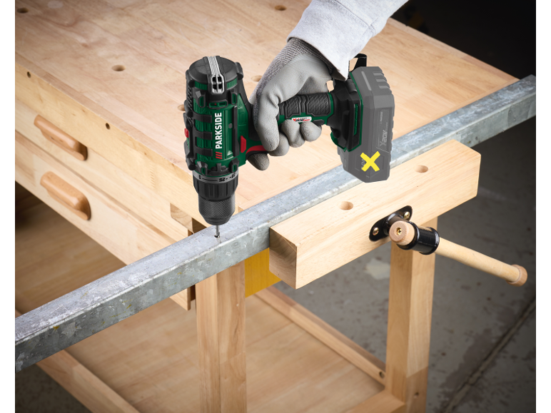 Green and black cordless drill drilling a metal bar on a wooden workbench.