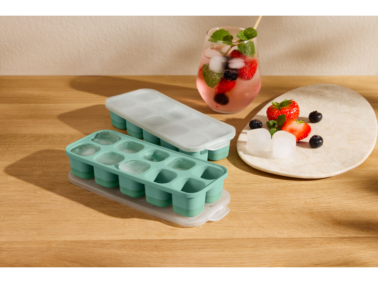 Two ice cube trays, one filled with ice, next to a berry drink and fresh fruit.