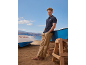 Man in navy polo shirt and khaki trousers on the beach, leaning against a boat.