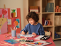 Child painting with watercolors and brushes on a pink table, with artwork in the background.