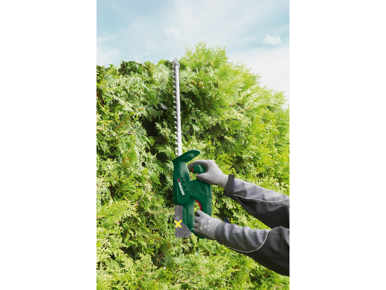 Green electric hedge trimmer cutting a cypress hedge under a blue sky.