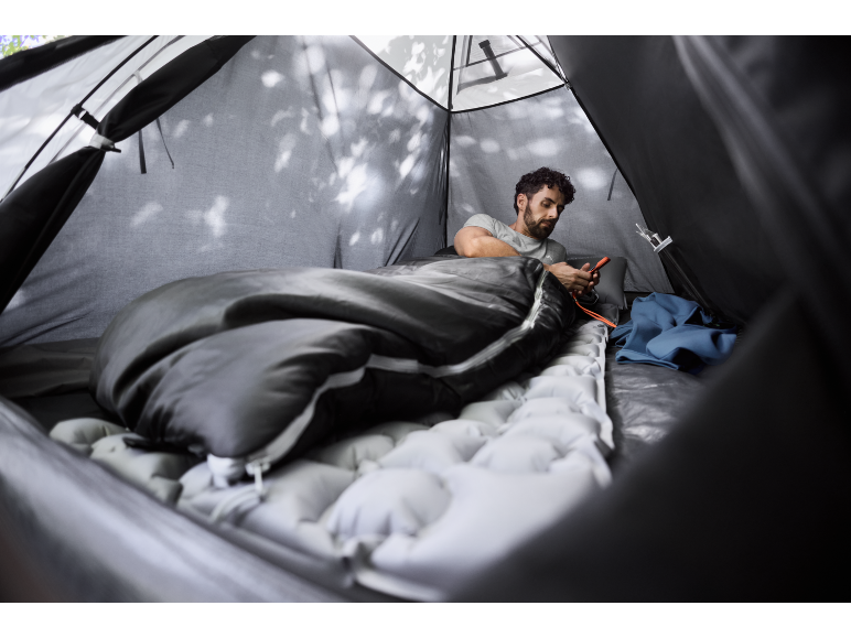 Man in a sleeping bag inside a tent, using a smartphone.