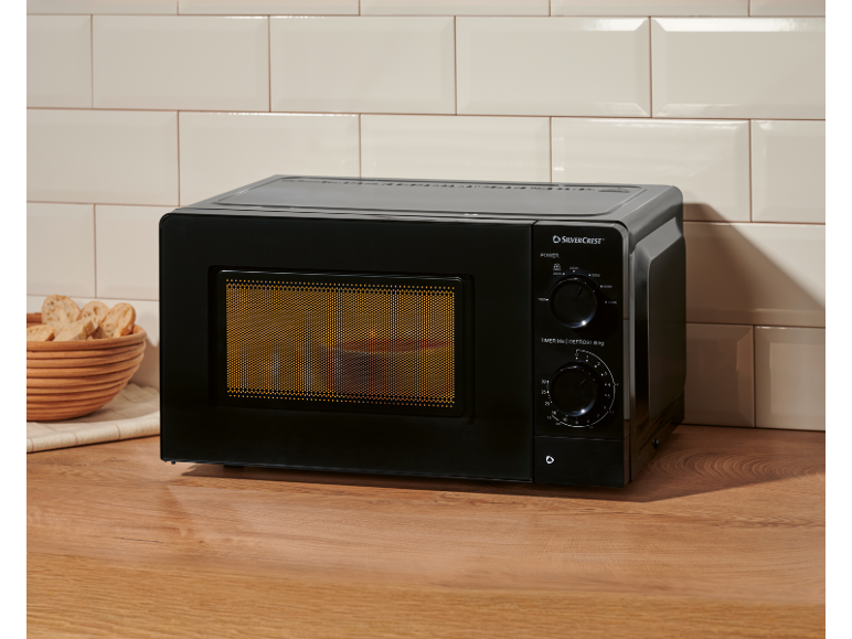 Black microwave oven on a wooden countertop with tiled background and a basket of bread.
