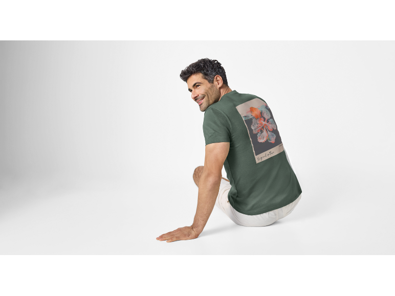 Man in a dark green t-shirt with a floral graphic on the back, sitting and smiling.