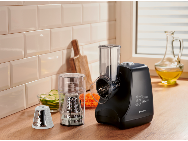 Black electric slicer with cutting and grating attachments, surrounded by fresh vegetables.