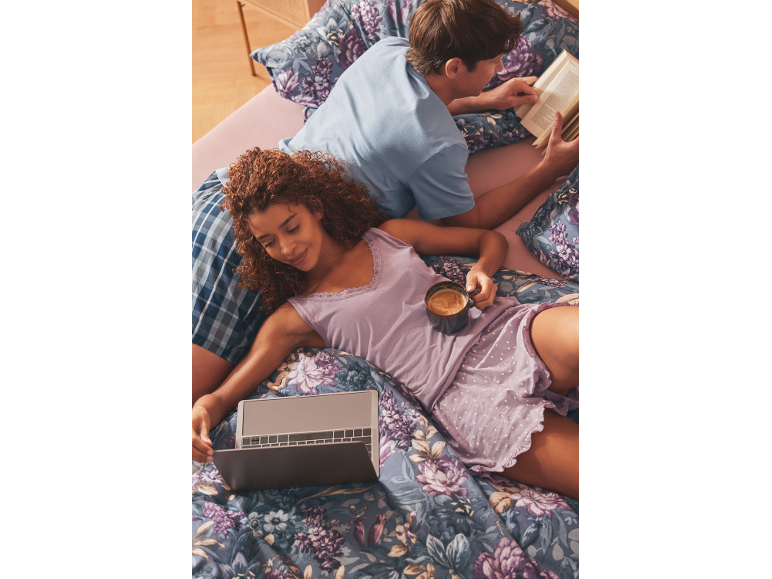 Couple in bed with floral bedding, woman with laptop and coffee, man reading a book.