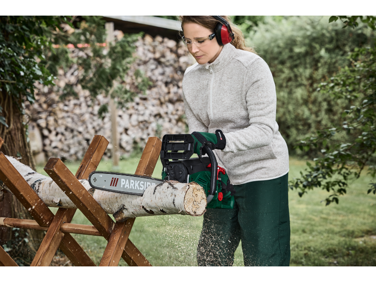 Woman with chainsaw cutting a wooden log on a sawhorse, wearing eye and ear protection.