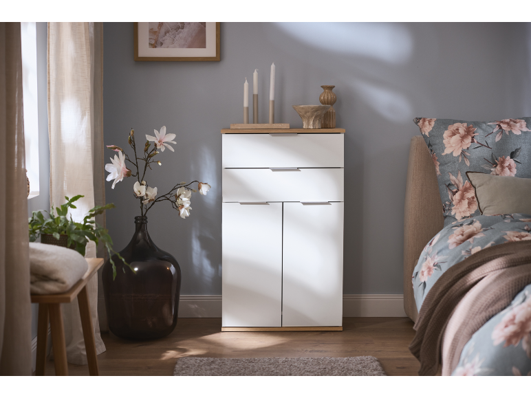 White cabinet with drawers and doors, decorated with flowers and candles, next to a bed.
