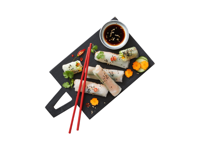 Fresh spring rolls with dipping sauce, red chopsticks, and garnishes on a dark board.