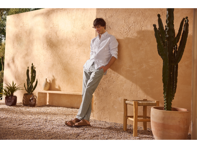 Man in white shirt and grey trousers, sandals, leaning against a sand-colored wall with cacti.