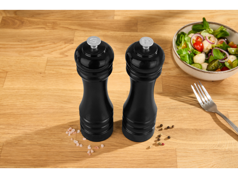 Black salt and pepper mills on a wooden table with salad and a fork.