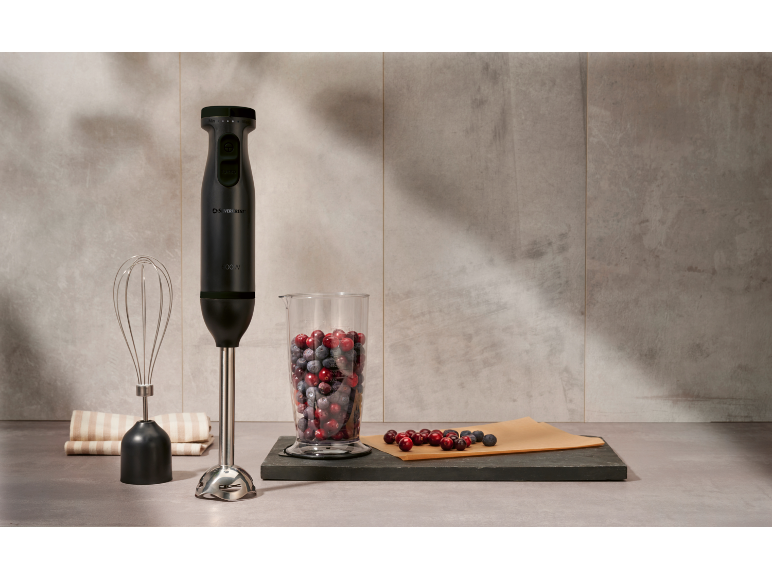 Black hand blender with attachments and berries in a measuring cup on a kitchen counter.