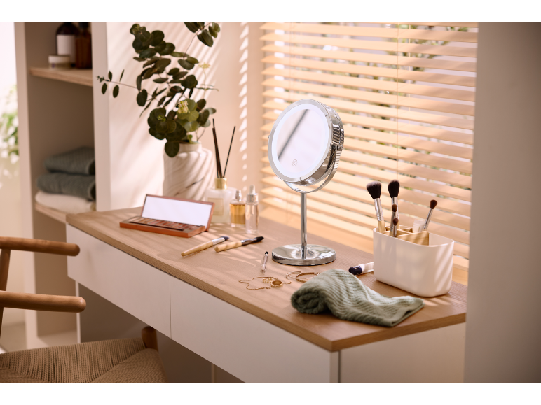 Dressing table with an illuminated mirror, cosmetics, and brushes, by a window with blinds.